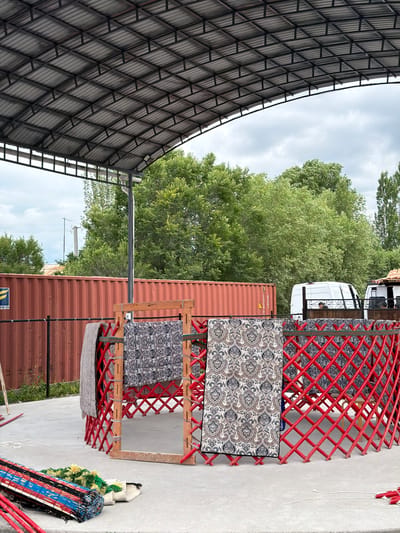 The image depicts an outdoor scene in the city of Кызыл-Туу, Kyrgyzstan, under a large, gray, corrugated metal roof supported by a dark metal frame. In the foreground, a partially constructed yurt-like structure with red lattice walls and wooden uprights is visible. Several patterned textiles are draped over the structure, including a gray blanket and two large, ornate fabrics. To the left, a rolled-up mat with red, blue, and black stripes lies on the concrete ground, along with some loose red poles and white rope. In the background, a long, red shipping container stands behind a black metal fence. Beyond the container, a line of lush green trees forms a backdrop against a cloudy sky. Two white vans are parked behind the yurt structure, with glimpses of people inside or near them. The overall impression is one of an outdoor gathering or construction area, possibly related to traditional dwelling setup or an event.