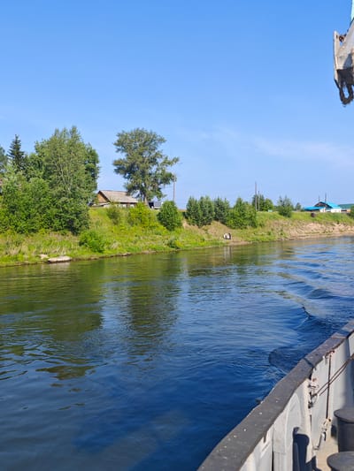 The image shows a wide river with a boat in the foreground, viewed from its starboard side. The river water is dark blue with rippling waves. On the opposite bank, a grassy slope rises to a treeline with several houses and structures. The sky is clear and blue. In the middle ground of the far bank, a small group of people, appearing to be two adults and one child, are standing near the water's edge. The scene depicts a rural or suburban setting along a river, potentially in or near the village of Galanino, Russia, given the context. The time of day appears to be daytime, likely late morning or afternoon, due to the bright sunlight.