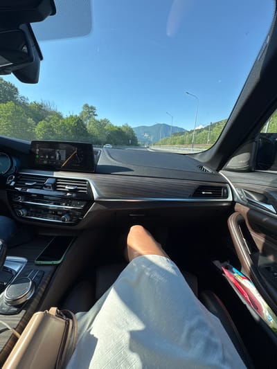 This is a point-of-view shot from inside a car, traveling on a highway. The dashboard is visible, featuring a large infotainment screen displaying music and the date "18.06.2026". Below the screen are climate control buttons and vents. The focus is on the interior of the car, with a person's legs and a handbag in the foreground. Outside the car, a bright blue sky, green trees, and mountains can be seen. The road ahead is clear with some distant vehicles. The specific location of Galitsyno, Russia, is not discernible from the visual information provided in the image.