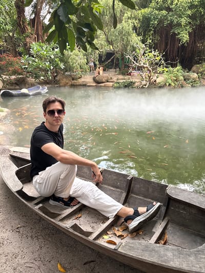 A man wearing sunglasses, a black T-shirt, and white pants sits in a weathered wooden boat. The boat is situated next to a pond teeming with orange fish. Vapor rises from the pond's surface. Lush vegetation surrounds the pond, including trees with exposed roots. The scene is located in the city of Nam Nha Trang, Vietnam.