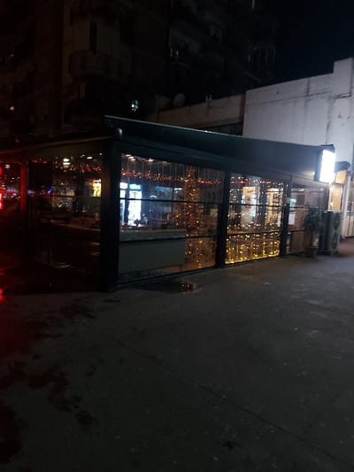 This night-time image, originally rotated ninety degrees counter-clockwise, depicts an urban street scene in Tiranë, Albania. The central focus is a modern, glass-fronted building, likely a shop or cafe, brightly lit from within.
The building's large windows display numerous warm-toned string lights (fairy lights) and reflections of the street and buildings opposite. The interior appears inviting, with glimpses of furniture and other elements, though specific details are obscured by reflections and distance. Above the entrance, a rectangular sign is illuminated, but any text or logo on it is illegible due to blurriness and angle.
To the left of the building, other dark, less distinct buildings line the street. The foreground on the right features a paved sidewalk or road, appearing dark and slightly textured. There's a subtle red glow at the bottom edge, possibly from a vehicle's tail lights or a distant neon sign, reflecting on the wet-looking surface.
The scene is quiet, with no people visibly present or discernible activities taking place. The overall atmosphere is calm, lit predominantly by the artificial lights of the building and reflections from the urban environment. The time of day is clearly night, and the weather appears clear.