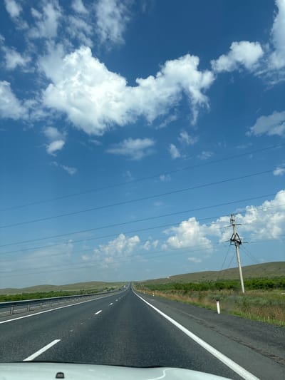 The image depicts a view from a vehicle driving on a paved highway under a bright blue sky with scattered white clouds. The road stretches into the distance, bordered by a metal guardrail on the left and a grassy, undulating landscape on the right. Power lines are visible overhead, supported by concrete poles. The terrain appears rural, with low hills covered in green vegetation and scattered trees. There are no people visible in the image, and no specific landmarks are identifiable that would indicate the location in Chernogorsk, Russia. The overall scene is one of travel on an open road in a natural environment.