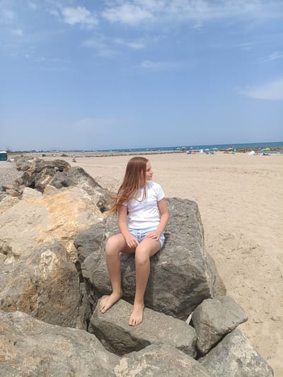 A young girl with long, auburn hair is sitting on large rocks by the sea. She is wearing a white t-shirt and blue shorts. She is looking to her right, away from the camera. Behind her is a sandy beach with many umbrellas and people. The sky is blue with some scattered clouds. The scene is located in Almardà, Spain.