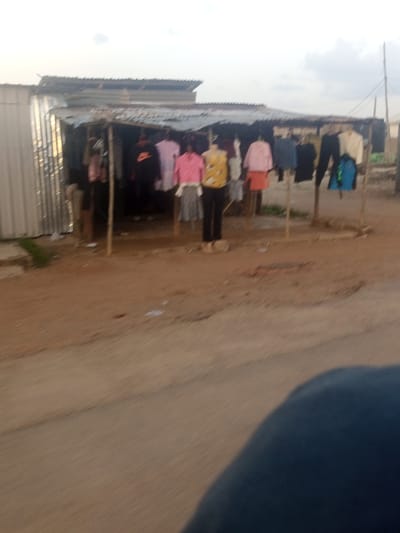 A street-level outdoor stall displays clothing on hangers and mannequins. The stall has a corrugated metal roof supported by wooden poles. Clothing items include shirts, dresses, and pants in various colors such as pink, yellow, blue, and black. Two mannequins are present, one wearing a yellow and black patterned top with black pants, and another wearing a pink top and an orange skirt. The stall is situated on a dirt ground adjacent to a paved road. A corrugated metal structure is visible to the left of the stall. The sky is overcast with scattered clouds. The image is taken from a moving vehicle, resulting in motion blur.