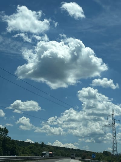 The image depicts a cloudy sky above a highway in the vicinity of Verinsko, Bulgaria. The sky is a vibrant blue, dotted with numerous white, fluffy cumulus clouds of varying sizes. Several power lines stretch horizontally across the frame, obscuring parts of the sky and landscape. On the right side of the image, a tall, metallic electricity pylon stands prominently against the clouds.
Below the sky, a tree-covered hillside forms the horizon. The highway is visible in the foreground, with a few vehicles including cars and trucks. A traffic sign with blue arrows indicating a diversion is also visible on the right side of the highway. The overall scene suggests a clear day with scattered clouds, likely captured from a moving vehicle on the highway.