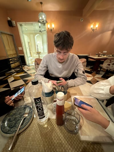 At a restaurant table, three people are occupied with their phones. One person wears a light grey shirt and glasses. Another is in white. A black and white checkered floor is visible. On the table, there's cutlery, a bottle of ketchup, a water bottle, a glass, and plates, including one with a fork. The table is topped with a woven placemat. The restaurant interior features lights and a view through an archway.