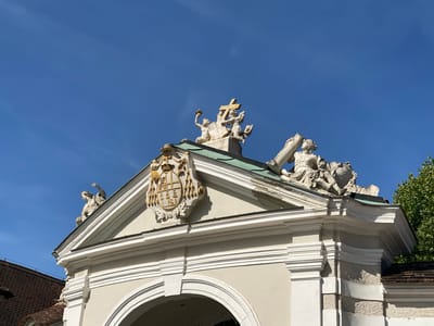 The image shows the top of an ornate gateway in Sankt Pölten, Austria. The gateway is topped with several classical sculptures against a clear blue sky. Prominently featured is a coat of arms in gold and yellow, located on the pediment of the gateway, which is topped with a green roof. Above the pediment, a cross is being held up by two cherubic figures. To the right, a larger sculpture depicts a female figure holding a large cylindrical object. To the left of the gateway's pediment, another sculpture of a seated figure is visible. The architectural style suggests a historical or ecclesiastical building.