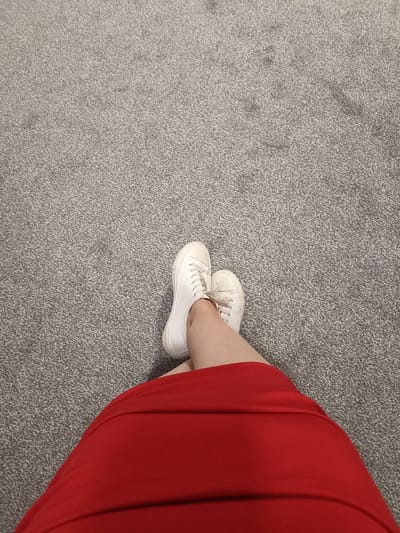 An overhead, eye-level shot shows a person's legs from the mid-thigh down, wearing a bright red skirt and white canvas sneakers. The person's legs are crossed at the ankles, and their feet are resting on a gray carpet. The carpet has a flecked texture and appears to be a standard indoor carpeting. The scene is devoid of any identifying landmarks, making it impossible to determine the specific location within Annagry, Ireland. There are no other people or objects visible in the frame. The lighting suggests an indoor setting, possibly a home or a public building.