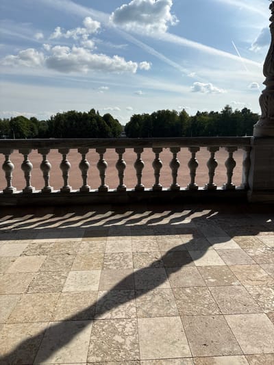 The image depicts a view from a stone-tiled balcony overlooking a wide, reddish-brown plaza. A decorative balustrade with ornate stone balusters runs horizontally across the middle ground, casting long, distinct shadows on the tiled floor in the foreground. Beyond the plaza, a dense line of green trees forms a backdrop, with a bright, partly cloudy sky above. The setting appears to be the Gatchina Palace in Gatchina, Russia, known for its expansive grounds and classical architecture. The light suggests it is daytime, likely late morning or early afternoon. There are no people visible in the frame.
