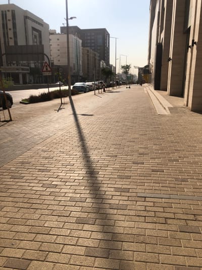 A street scene unfolds with a brick-paved sidewalk extending into the distance. Tall buildings line the left side of the frame, casting shadows onto parked cars. A street light's shadow stretches across the bricks. A road sign is visible near a strip of green vegetation separating the sidewalk from the street. The sky is a pale blue, suggesting a sunny day.