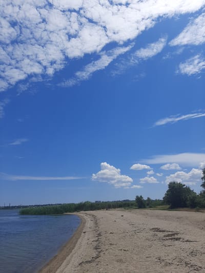 A bright, sunny day is captured in this landscape photograph. The scene is divided into a clear blue sky with scattered white clouds and a tranquil body of water meeting a sandy shore. The water is calm, with gentle ripples reflecting the sky. The sandy beach appears dry and stretches along the edge of the water.

Behind the beach, a dense line of green reeds or tall grass grows, forming a natural border. Further back, a line of trees with dark green foliage can be seen, suggesting a natural or park-like setting. In the distance, faint structures or buildings are visible, hinting at a populated area or a town nearby.

The time of day appears to be midday, indicated by the bright sunlight and the vivid blue of the sky. The weather is clear and likely warm, suitable for outdoor activities. The location is identified as Sukholuzhzhia, Ukraine, which, combined with the presence of water and greenery, could imply a lakeside or riverside environment.

The image evokes a sense of peace and natural beauty. There are no people or specific objects of focus, drawing attention to the serene landscape itself. The composition, with the sky occupying a significant portion of the frame, emphasizes the vastness of the heavens and the airy atmosphere of the day. The orientation of the photo is landscape.