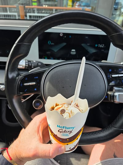 A person's hand is holding a McFlurry in a paper cup in front of a car's steering wheel. The McFlurry appears to be a Galaxy Salted Caramel flavor, with ice cream, chocolate pieces, and caramel sauce. The steering wheel has control buttons and a digital display screen visible in the background. The car is likely parked in Milton Keynes, United Kingdom, given the presence of the specific McDonald's product and the context of the vehicle. The overall scene suggests a casual moment of enjoying a treat while in a car.
