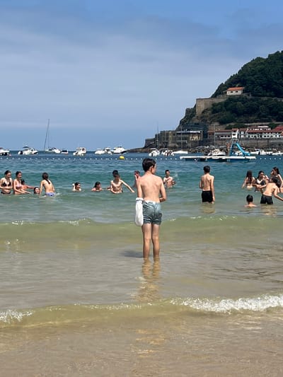 A group of people, primarily young individuals, are enjoying a sunny day at La Concha Beach in San Sebastián, Spain. The scene captures the vibrant atmosphere of the popular tourist destination. In the foreground, a young boy stands facing away from the camera, partially submerged in the shallow, clear, turquoise water. His back is to the viewer, and he wears light-colored shorts and has a white bag slung over his shoulder. Behind him, a diverse group of people are scattered throughout the water. Some are interacting with each other, while others are simply swimming or wading. The water is calm and inviting, with small waves gently lapping at the shore.

In the mid-ground, several boats are anchored in the bay, their white hulls and masts creating a picturesque backdrop. Beyond the boats, the distinctive coastline of San Sebastián is visible. The lush green slopes of Monte Urgull rise dramatically from the sea, topped with a historic fortress. The buildings of the city are nestled along the hillside and at the base of the mountain, painted in various shades of white, cream, and terracotta, adding to the charming aesthetic of the location. The sky is a clear, bright blue, indicating a perfect summer day for beach activities. The overall impression is one of leisure and enjoyment in a beautiful coastal city.
