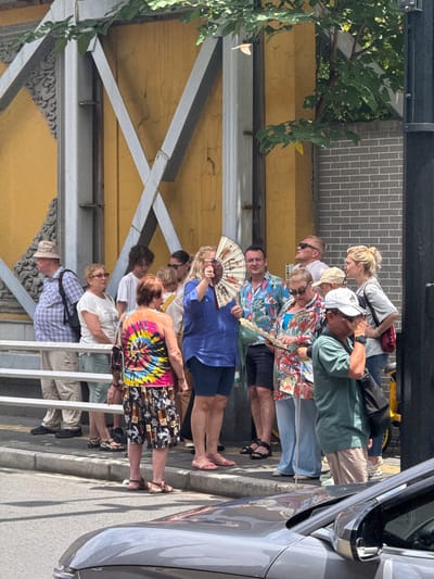 The image captures a daytime scene in what appears to be an urban environment, likely in the Jing'an District, China. A group of approximately a dozen people, ranging in age, are standing in a line or queue on a paved walkway.
The individuals are dressed in casual summer clothing, including shorts, t-shirts, light blouses, and sandals, suggesting warm or hot weather. Several people are holding or actively using fans to cool themselves; one person in a blue top is seen fanning themselves with a decorative, traditional Chinese-style fan adorned with painted figures. Other notable attire includes a brightly colored tie-dye top and patterned black shorts with white anchor motifs and the word "SAILOR" repeated. Many wear sunglasses.
To the right, the front fender and headlight area of a silver or grey car are visible, parked alongside the curb. The background features a building with a textured yellow wall, showing signs of wear and cracking, and above it, a section of a grey brick wall with white mortar. Green foliage from trees or vines cascades down from above the structures on the left side of the image. Metal beams and decorative architectural elements are also present, forming part of an overhead structure or building facade. The bright lighting and distinct shadows indicate it is daytime. The people appear patient, and the overall mood is calm.