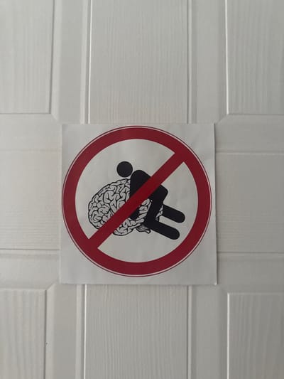 A circular prohibition sign with a red border and a red diagonal line across it is adhered to a white, paneled door. Inside the circle, a black silhouette of a person appears to be straddling or riding a black, stylized representation of a human brain. The person's head is a solid circle, and their body is depicted in a leaning, forward posture. The brain graphic is detailed, showing convolutions. The overall impression of the sign is a humorous or cautionary message against using one's brain, or perhaps against "brain-dead" actions. The door appears to be an interior door, possibly in a residential or office setting. There is no discernible indication of the specific location within Dublin, Ireland, as the image only shows the sign and the door.