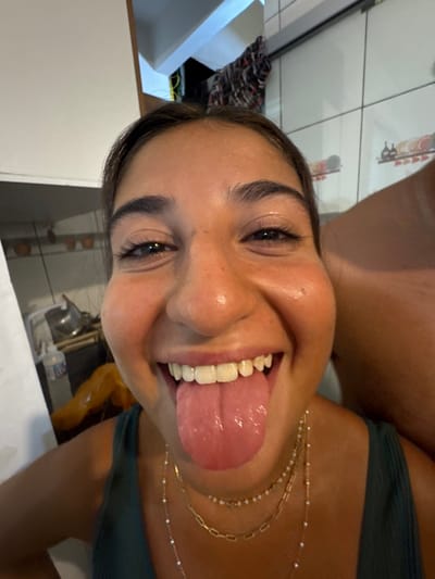 A woman smiles, sticking out her tongue. She has dark hair and eyebrows, light skin, and wears a green top. Three delicate gold necklaces adorn her neck. The background includes a tiled wall, a white countertop, and a glimpse of a kitchen sink and cabinets. The overall perspective is from a slightly low angle.