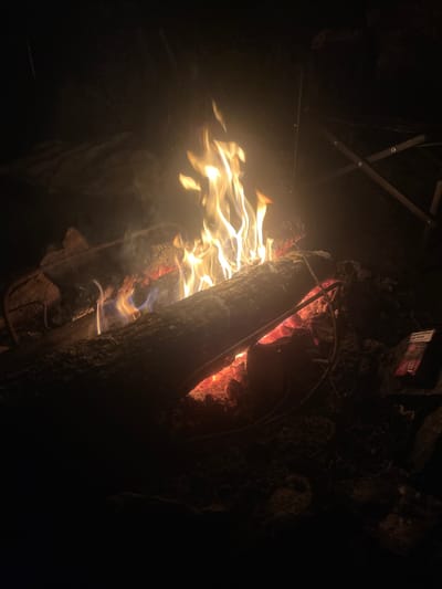 A campfire burns in the dark. Flames flicker and dance above logs and embers, casting a warm glow on the surrounding ground. The scene is set against a black background, suggesting nighttime or a dimly lit area. A metal folding chair is visible in the upper right corner, partially obscured by smoke and shadows. In the lower right, a red and white package, possibly of cigarettes, lies on the ground. The overall atmosphere is one of a natural, outdoor setting, possibly a campsite or a gathering around a fire. The location in Mala Kokhnivka, Ukraine, is not visually evident from the image itself.