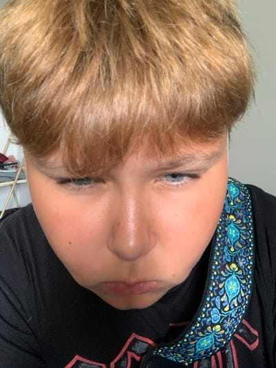 This media file is a close-up, tilted photograph of a young person's face. The subject has light brown hair and blue eyes. They are wearing a black shirt with a decorative collar featuring a blue and yellow embroidered pattern. The image is well-lit, suggesting it was taken indoors during the daytime. The person's expression is neutral. No text is visible in the image. The location context provided is Vejprty, Czechia.