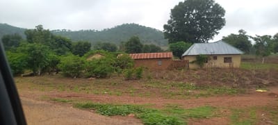 The image depicts a rural landscape in Gu, Nigeria, characterized by a cloudy sky and a lush, hilly terrain covered in trees and vegetation. In the foreground, a dirt road with patches of green grass runs horizontally across the frame. Behind the road, there is a clearing with scattered dry plants and soil. Two buildings are visible in the middle ground. The building on the left is a small, light-colored structure with a flat roof. To its right, a red-tiled roof building stands, appearing to be constructed from earth or clay. Further to the right, a larger building with a silver metal roof is partially visible, nestled amongst trees. The background is dominated by a large, dark green tree and a distant, mist-covered hill. The scene appears to be a residential area in a village or outskirts of a town.