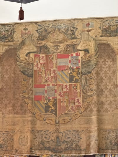 This is a detail of a large, antique tapestry, likely displayed indoors. The tapestry features a prominent coat of arms, intricately woven with various symbols and heraldic elements. The central shield is quartered, displaying a lion rampant in one section and checkered patterns of red, white, and blue in others, alongside depictions of castles and more lions. The coat of arms is flanked by elaborate decorative motifs, including what appear to be stylized wings or feathers, and ornate scrolling patterns typical of historical tapestries. The background of the tapestry is a repeating pattern of fleur-de-lis. The overall color palette is muted, with shades of brown, beige, red, blue, and gold, suggesting age and a historical context. In the lower left of the image, a portion of a white wall is visible, and a dark brown tassel hangs from a thin cord, possibly part of a curtain or window dressing. The tapestry's condition appears somewhat worn, with visible creases and some fraying at the edges. The lighting suggests an indoor setting, with the light source casting subtle shadows on the textured surface of the tapestry. No discernible text is visible. Given the location context of Sevilla, Spain, this tapestry might be part of the historical collection of a palace, museum, or significant building in the city, potentially depicting the lineage or authority of a historical figure or entity.