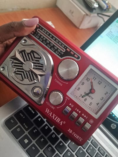 A red "WAXIBA® XB-742BTC" radio, a model with an analog clock. The clock face shows the time around 6:50. The radio includes tuning and volume knobs and multiple playback buttons. Frequency bands in MHz, KHz, and MHz are marked on the radio. The radio sits on a silver laptop, partially obscuring the keyboard. A hand with painted nails is holding the radio. A computer monitor and office supplies are visible in the background.