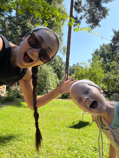 This media file features a selfie taken outdoors, likely in Udelnaya, Russia, given the provided context. Two subjects are visible: a woman in the foreground and a child in the background, both appearing to be taking the photo together. The woman is wearing sunglasses and a black top, with a white bandana on her head and her long, dark hair braided and extended outwards. She has an open-mouthed expression, conveying excitement or surprise. The child, positioned upside down relative to the woman, is also looking at the camera with a wide, open-mouthed expression, wearing a light-colored garment and a white bandana. The setting is a sunny outdoor environment with lush green grass and trees, suggesting it's daytime with pleasant weather. A thin wooden pole and some hanging string lights are visible in the upper left portion of the frame, adding to the outdoor ambiance. The overall mood is playful and joyful, capturing a moment of shared fun between the two individuals.
