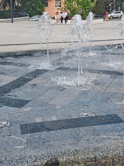 The image depicts a ground-level fountain with multiple water jets spraying upwards on a paved square in Noginsk, Russia. The paving consists of gray granite-like tiles with darker, rectangular insets. Water pools on the tiles between the jets, some of which have drains at their base. In the background, two people are seated on a low concrete wall. A child in a pink hat walks away from the scene. Several cars are parked in the vicinity, and a building with signage can be seen behind the people. Trees are present, suggesting a park or urban green space.