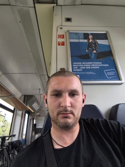This is a close-up, selfie-style photo taken inside a train. The main subject is a man with short brown hair and a beard, wearing a black t-shirt and black earbuds. He is looking directly at the camera with a neutral expression.

Behind the man, the interior of the train is visible. To his left, a window shows a blurry outdoor scene with greenery and a bicycle rack. There are also two bicycles visible near the window. The train carriage appears to be modern, with beige walls, overhead storage, and dark blue seats.

On the wall above and behind the man's head, there is a large poster. The poster features a smiling woman wearing a floral jacket and jeans, standing against a blue background. Text on the poster is in German. The visible text includes: "UNSERE MITARBEITENDEN MACHEN EINEN GROSSARTIGEN JOB - UND WIR SAGEN: DANKE!" which translates to "OUR EMPLOYEES MAKE A GREAT JOB - AND WE SAY: THANK YOU!". Below this, there is smaller text that appears to be an advertisement or company information. In the top right corner of the poster, a smaller box of text says "WIR sind deine Mobilitätspartnerin in der Region" which translates to "WE are your mobility partner in the region". Further text at the bottom of the poster indicates the company name as "Bäderbahn GmbH" and provides an address.

The lighting suggests it is daytime, and the outdoor scene through the window does not indicate specific weather conditions. The man is seated, possibly on his commute or traveling. The setting is clearly a public transportation vehicle.