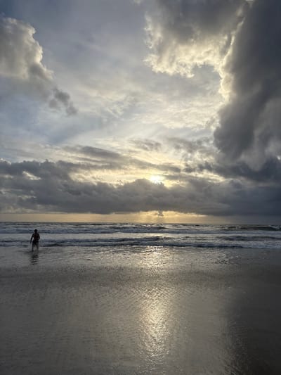 A beach scene shows dark storm clouds with light breaking through. Waves gently roll onto the wet sand, reflecting sunlight. A lone figure wades in the water near the shoreline. The sky dominates the image with its dramatic cloud formation. The beach is mostly smooth with gentle ripples from the receding waves. The overall tone is serene, yet with a hint of the power of nature.