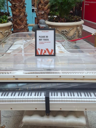 The image shows a clear acrylic grand piano in what appears to be a public space, possibly a shopping mall or atrium, given the presence of potted plants and the general decor. A sign is prominently displayed on the piano stating "PLEASE DO NOT TOUCH. This piano is finely tuned and very delicate." The piano itself has white and black keys, and its transparent casing reveals the intricate internal mechanisms. To the left, a person wearing a grey baseball cap and a dark t-shirt is partially visible, appearing to be looking at the piano or the sign. In the background, a red wall with text indicating "Until then, find us upstairs at our usual location" suggests a commercial establishment. The overall lighting suggests daytime. The location is identified as Manchester, United Kingdom.