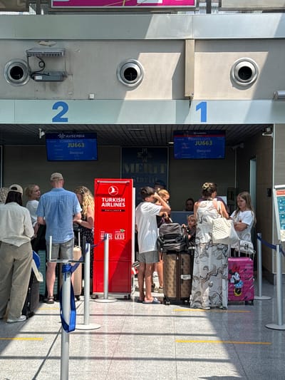 This is a vertical, full-shot photograph taken outdoors at what appears to be an airport. The sun is shining, casting strong shadows on the grey tiled ground, suggesting it is daytime. The scene depicts a queue of people, likely passengers, waiting in line. They are standing next to various pieces of luggage, including suitcases and backpacks, some of which are quite colorful and patterned.

Two prominent blue information screens are visible. One displays "AirSerbia BUSINESS CLASS JU663 Beograd 15:15" with a time of "13:53". Another screen shows "AirSerbia ECONOMY CLASS JU663 Beograd 15:15" also with the time "13:53". A red stand with "TURKISH AIRLINES STAR ALLIANCE" branding is also present, featuring text about lighter luggage.

The people in the queue are casually dressed, some wearing t-shirts, shorts, and hats. Their expressions are neutral or slightly focused, indicative of the mundane activity of waiting for a flight. The overall atmosphere is one of travel and transit. The presence of numbered signs ("1" and "2") suggests this might be a boarding gate or check-in area. The location context provided is Golubovci, Montenegro, which aligns with the presence of AirSerbia, the national airline of Serbia, which operates flights to and from Montenegro.