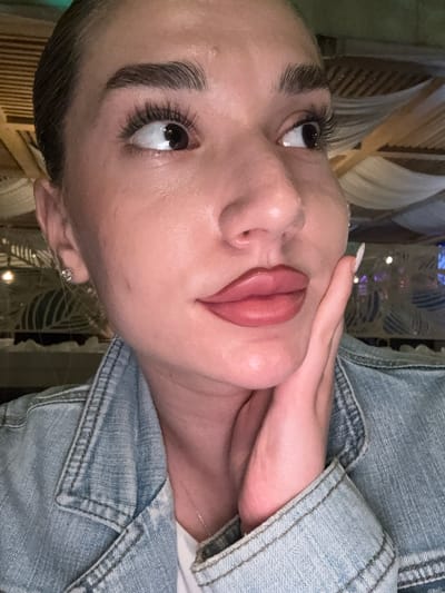 The image is a close-up, vertically oriented portrait of a person's face, with the camera angled downwards. The subject, who appears to be a woman, has her face turned to the side, with her right hand gently touching her face near her chin. She is wearing a light blue denim jacket. Her makeup includes mascara on her eyelashes and a matte, rosy-toned lipstick. She is also wearing a small, diamond stud earring in her visible ear. The background is blurred, but appears to be an indoor setting with decorative elements, possibly curtains or a patterned screen, and some warm lighting. The lighting suggests it might be evening or a dimly lit indoor environment. There is no visible text in the image. The overall mood of the image is intimate and personal, focusing on the subject's features and makeup.