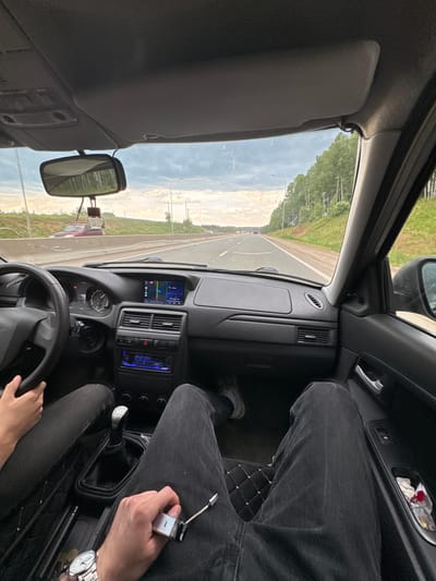 The image shows the interior of a car driving on a highway, with a view of the road ahead and the surrounding landscape. The perspective is from the passenger seat, looking towards the driver. Two individuals are partially visible: the driver's hands are on the steering wheel, and the passenger's legs are in the foreground, with one hand holding a car key and the other resting on their lap. The car's dashboard, infotainment system, gear stick, and interior door panel are visible. The road appears to be a multi-lane highway, with trees and vegetation lining the sides. The sky is cloudy. The specific location is the city of Beloborodovo, Russia.