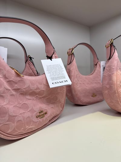 Here is a description of the image:

The image displays three pink Coach handbags, stacked on a shelf. The handbags are the same style, with a curved shape and a short strap. They are made of a fabric with a Coach logo pattern. Each bag has a tag attached. The tags have text in both English and French, including information about the materials used and the brand. The overall setting appears to be a retail environment. The location is in the city of Sunrise, United States of America.