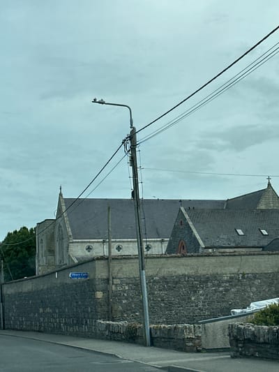 The image captures a cloudy day scene in the city of Carlow, Ireland. Dominating the foreground is a street light with several power lines running across the frame. To the left of the street light, a sign on a stone wall reads "Sleaty Cafe". Behind the wall, the roofline of a church is visible, with a cross atop its spire. The church is constructed of grey stone and features arched windows. Several birds are perched on the roof of the church and on the street light. The overall environment suggests a quiet, possibly overcast, day in a built-up area.