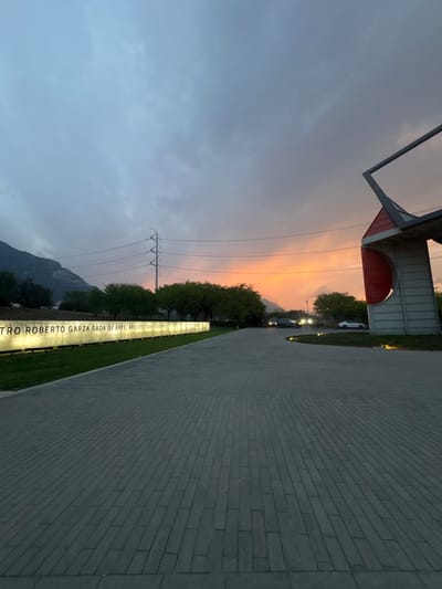 The image captures an outdoor scene at dusk in San Pedro Garza García, Monterrey, Mexico, facing east towards the Centro Roberto Garza Sada at the Universidad de Monterrey (UDEM). A wide, gray, interlocking paver roadway dominates the foreground, receding into the distance. To the left, a long, illuminated sign displays the text "CENTRO ROBERTO GARZA SADA DE ARTE, ARQUITECTURA Y DISEÑO." Beyond the sign, a dark mountain range is visible on the horizon beneath a gray-blue, cloudy sky. An electricity pylon with multiple power lines stands in the mid-ground. The lower right portion of the sky is illuminated by an orange and yellow sunset. Green trees line the roadway and partially obscure the horizon. In the mid-ground, a curved asphalt road contains at least two dark vehicles with illuminated headlights and a parked white vehicle. On the far right, a modern architectural structure featuring red panels, gray components, and an upward-angled rectangular frame is partially visible. No people are present.