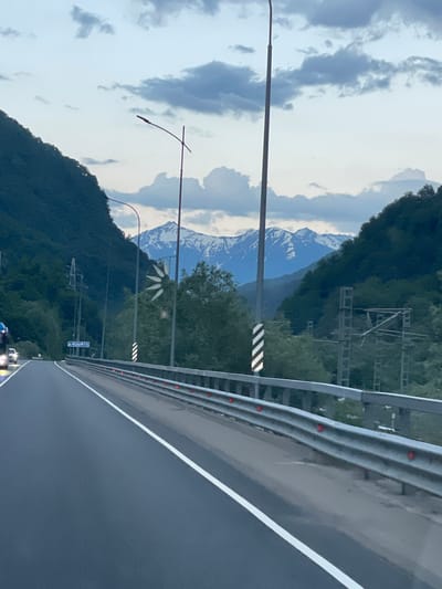 The image shows a road curving through a mountainous landscape, likely at dusk or dawn given the soft lighting and cloudy sky. The road is paved and has a white lane marking. A metal guardrail runs along the side of the road, with red reflectors. On the left side of the road, a steep, tree-covered mountainside rises up. On the right side, there are more trees, overhead power lines, and a distant view of snow-capped mountains. A signpost with blue text is visible on the left side of the road, indicating the location as "р. МЗЫМТА" (River Mzymta). The image appears to be taken from a moving vehicle.