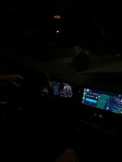 The image depicts the interior of a vehicle at night, viewed from the passenger side, facing the dashboard. A driver's left hand is visible on the steering wheel. A second hand is visible in the lower right corner, likely belonging to a passenger.
The digital instrument cluster behind the steering wheel displays a map segment and "-21.0 C" data. A prominent central infotainment screen shows a navigation application, likely Apple CarPlay, with a map of an urban area. Street names visible on the map include "R. de Mouche", "R. de Mar. Saldanha", and "R. de Pedro de Covilhã". A Portuguese language pop-up on the navigation screen asks, "Conduzir até casa?" (Drive home?), offering "SIM" (Yes) and "NÃO" (No) options, with a route estimated at "7 min - 1.6 km." Below the infotainment screen, vehicle controls with red and orange indicators are partially visible. A circular, dark air freshener or ornament, possibly with "SCX" text, hangs from the rearview mirror area. An illuminated "PASSENGER AIR BAG OFF" indicator is visible on the upper dashboard. The external environment is dark, with minimal ambient light suggesting night conditions.
Based on the visible street names on the navigation display ("R. de Mouche", "R. de Mar. Saldanha"), the vehicle is located in the city of Porto, Portugal, specifically within the civil parish of Aldoar, Foz do Douro e Nevogilde.