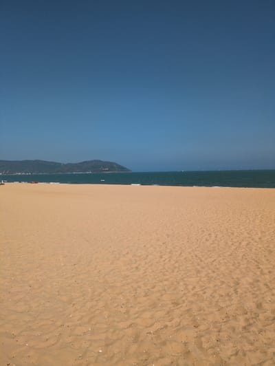 The image depicts a view from above of a sandy beach adjacent to the ocean in the city of Quy Nhon Ward, Vietnam. The horizon is a vertical line between the tan-colored beach and the dark blue sky. Between the sky and the sand is a line of water. A large, rocky hill rises along the shoreline of the water. The beach shows a pattern of footprints and scattered debris.