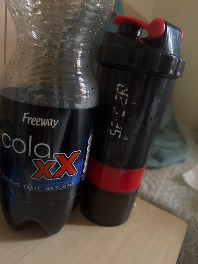 The scene depicts an indoor environment in Alderbury, United Kingdom. Two beverage containers stand upright on a light-colored wooden surface. To the left is a clear plastic bottle partially filled with a dark liquid, identified by its label as "Freeway cola XX Extreme Taste, No Sugar." Condensation droplets are visible on the exterior surface of this bottle. To the right is a black protein shaker bottle featuring a red flip-top drinking spout and a red band around its lower body. Volume markings, including "500 ML," are present on the side of the shaker, which also exhibits condensation droplets. The background consists of blurred textile elements, including a light-patterned fabric and a teal-colored object, alongside a plain light-colored surface. No individuals are present.