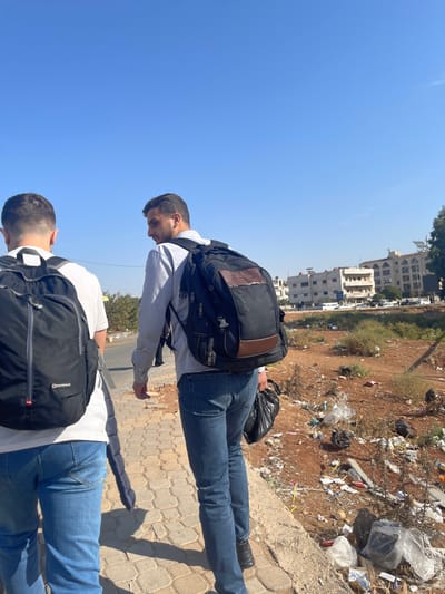 Two young men stand outdoors, each wearing a backpack and blue jeans. One wears a light-colored shirt, the other a white shirt with horizontal stripes. The camera angle is low, emphasizing the height and a view of buildings in the distance. Litter and foliage scatter across the ground in the background. The sky is clear and blue.