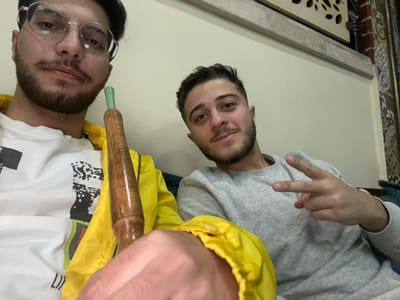 This image depicts two young adult males in an indoor setting within the city of Homs, Syria. The male in the left foreground, who appears to be taking the photograph, wears round clear-framed eyeglasses, a white long-sleeved shirt featuring a black and white graphic print, and a bright yellow jacket. He has dark hair, a mustache, and a beard, and his gaze is directed towards the camera. He holds a wooden hookah hose mouthpiece with a green tip in his left hand. To his right, the second male is seated, wearing a grey long-sleeved top. He has short dark hair and a beard, and he smiles at the camera while making a 'V' gesture with his right hand. The background consists of light-colored walls, with decorative elements visible on the right, including a panel with intricate patterns, possibly latticework, and a portion of patterned wallpaper. A dark green or teal colored cushion or upholstery is partially visible behind the second male.