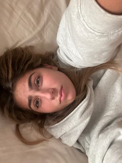 A young adult female is depicted lying supine on a light-colored, rumpled surface, likely bedding, in an indoor environment. The precise location within Aldoar, Foz do Douro e Nevogilde, Portugal, cannot be visually determined from the image. She is positioned diagonally across the frame, with her head in the lower-left quadrant and her torso extending towards the upper-right. Her head is slightly tilted, and she is looking directly towards the viewer. She has medium brown hair, fair skin with a slight flush on her cheeks, light-colored eyes, and prominent dark eyebrows. A small, thin hoop earring is visible in her left ear. She is wearing a light grey hooded sweatshirt. The lighting is soft and even, originating from an unidentifiable source. No other subjects or objects are visible in the plain background. No overt actions or interactions are occurring beyond the individual resting.