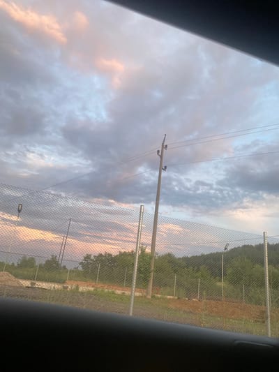 The image depicts a cloudy sky with patches of pink and pale blue hues, suggesting either sunrise or sunset. In the foreground, a chain-link fence stretches horizontally across the frame. Behind the fence, a landscape unfolds with trees and hills in the distance. A wooden utility pole with power lines is visible on the right side of the frame. There are also what appear to be streetlights or security lights mounted on poles scattered throughout the scene. The ground in front of the fence seems to be unpaved, with some areas of dirt and patchy grass. The overall impression is an outdoor scene, possibly on the outskirts of a town, given the combination of utility infrastructure and natural landscape. No people are visible in the image. The vantage point appears to be from inside a vehicle, indicated by the dark, blurry foreground element.