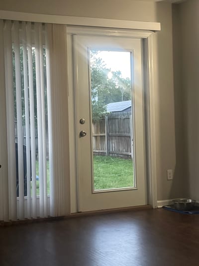 An interior shot shows a closed white-framed glass door with a doorknob and a deadbolt. To the left of the door, vertical blinds cover a window. Through the glass door, a wooden fence and green grass are visible in the backyard, with trees and a shed in the background. To the right of the door, a light-colored wall features a white electrical outlet. On the floor, a dark wood laminate is visible, with a metal pet bowl on a blue mat in the lower right corner. The scene is static and devoid of human presence. The image is not sufficient to determine the city of Jacksonville, United States of America.