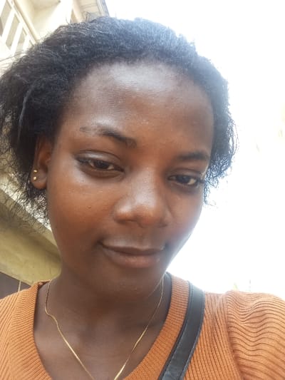 A close-up, head-and-shoulders photograph features a female individual with dark skin, dark, tightly curled hair, and dark eyes. She is positioned facing the camera, with a subtle smile and a slight downward gaze. Two small gold-colored stud earrings are visible in her left ear. A thin, gold-colored chain necklace rests around her neck. She wears an orange-rust colored, ribbed long-sleeved top. A dark strap, possibly from a bag, crosses over her right shoulder. The background consists of a light-colored building with a faint architectural detail visible behind her upper left side, and an overexposed bright white area occupying the right side of the frame, indicating strong daylight conditions. This scene is documented in Agburu Uke, Nigeria.