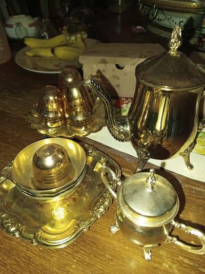 The scene depicts a collection of golden-colored metallic tableware on a dark wooden surface, presumably indoors, in Longmiang, Laos. In the immediate foreground, to the left, is an ornate, rectangular golden tray holding two stacked, small golden bowls, with an even smaller inverted golden cup resting atop the stack. To the right, a small, lidded golden container with a handle and three legs, resembling a sugar bowl, is positioned. Centrally, a prominent golden teapot with a curved spout, handle, and three decorative legs stands. Its lid features a detailed finial. To the left of the teapot, on a smaller, scalloped golden tray, are three smaller golden shakers, two cylindrical with ribbed bases and one spherical. In the background, on a white plate, are several ripe yellow bananas. Further behind, a partially visible white teapot with red markings is present. A light brown cardboard box is situated behind the golden tableware. The lighting is subdued, creating reflections on the metallic surfaces and casting soft shadows. No individuals are present. The objects are static, with no discernible actions or interactions occurring.