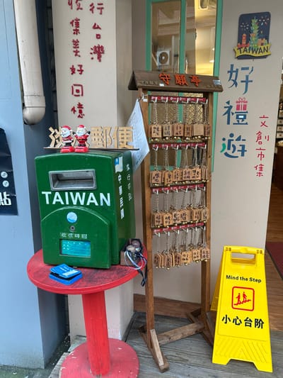 The image shows a collection of items next to a wall with Taiwanese characters and images. A green Taiwanese mailbox is prominently displayed, adorned with stickers and a Santa Claus figurine. Above it, wooden plaques hang from a rack. A yellow "Mind the Step" sign is placed to the right of the rack. The background wall is decorated with Taiwanese text and a Christmas tree image. Red letters and a pipe are seen along the wall.