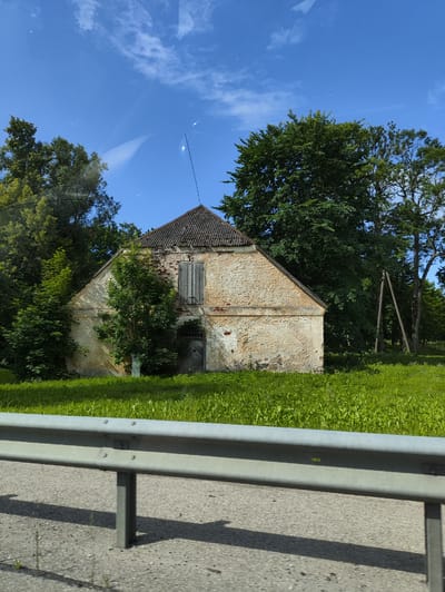 The image shows an old, abandoned building with a weathered stone facade and a decaying tiled roof, located in the rural area of Svētciems, Latvia. The building has wooden doors and shutters, and is surrounded by lush green grass and trees. In the foreground, a metal guardrail and asphalt road are visible, suggesting the building is situated near a road. The sky is a clear blue with a few scattered clouds, indicating a sunny day. There are no people or animals visible in the image. The overall impression is one of a neglected historical structure in a tranquil natural setting.