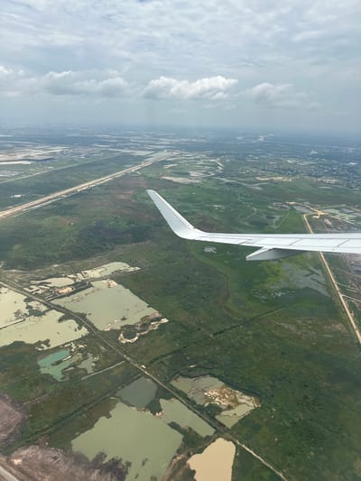 An aerial view shows the outskirts of Ta Khmau, Cambodia, with an airplane wing visible in the foreground. The landscape below is a mosaic of agricultural ponds and fields, with lush green vegetation dominating the scene. A straight, paved road runs through the agricultural area, connecting it to more distant urban areas that are hazy in the background. The sky is cloudy, with patches of blue peeking through. The overall impression is one of a rural or semi-rural area undergoing development, with a mix of natural and man-made features.